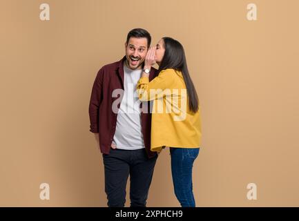 Jeune petite amie chuchotant secret à l'oreille d'un petit ami choqué. Couple habillé dans des commérages décontractés tout en se tenant ensemble isolé contre bac beige Banque D'Images