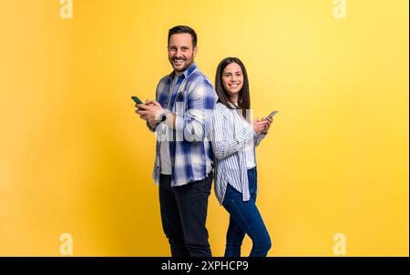 Portrait d'un couple souriant habillé en décontractés à l'aide de téléphones intelligents sur fond jaune. Joyeux jeune homme et femme debout dos à dos et moi Banque D'Images