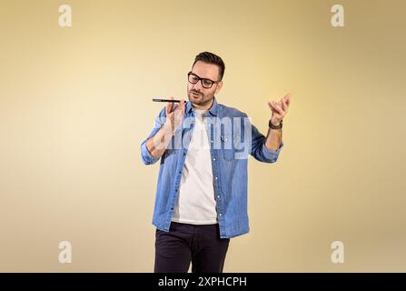 Portrait d'homme d'affaires habillé dans la chemise en denim envoyant la messagerie vocale par téléphone portable. Jeune homme parlant au téléphone portable et faisant des gestes en se tenant debout sur l'est Banque D'Images