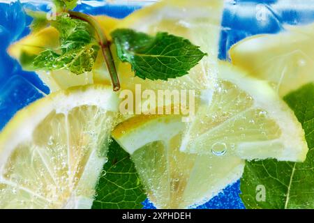 Des tranches de citron frais et des feuilles de menthe flottent dans de l'eau pétillante sur fond bleu. Papier peint texturé. Banque D'Images