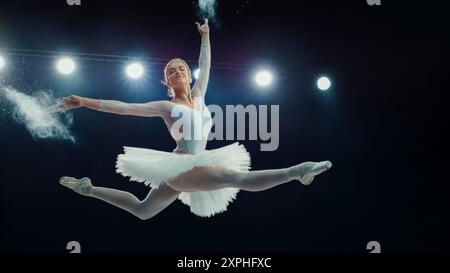 Gracieux Ballet dansant Ballerina en Tutu blanc interprète Dynamic Grand Jete, avec des lumières de scène illuminant son saut élégant et la poudre créant un effet dramatique, son poise et athlétisme. Banque D'Images