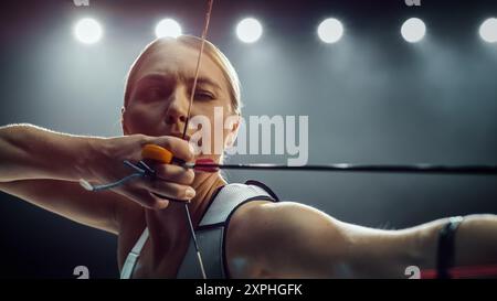 Portrait d'une belle Archer féminine tenant un arc composé moderne, visant une cible. Jeune athlète dessinant la flèche dans une compétition de stade ou dans un studio sombre pour la publicité Banque D'Images