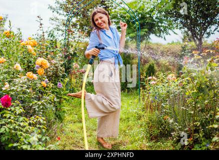 Femme heureuse jardinier ayant plaisir à arroser les plantes avec tuyau de tuyau dans le jardin d'été. Sauter sous des éclaboussures d'eau en fleurissant des roses debout sur l'herbe. Banque D'Images