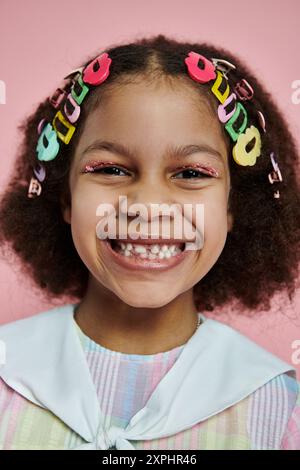 Une jeune fille afro-américaine aux cheveux bouclés ornée de pinces à cheveux colorées sourit brillamment sur un fond rose. Banque D'Images