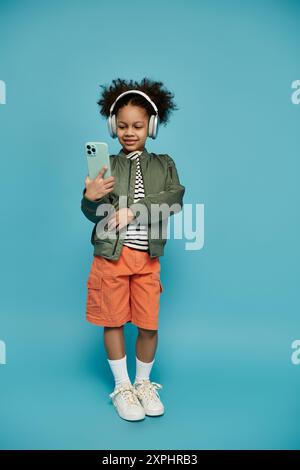 Une jeune fille afro-américaine aux cheveux bouclés est engrossée dans son smartphone, écouteurs sur fond bleu vif. Banque D'Images