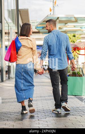 Vue arrière verticale de jeune couple faisant du shopping dans un centre commercial, portant des sacs en papier dans leurs mains et marchant ensemble se regardant et souriant. Mari et femme visitant la ville se tenant la main appréciant. Photo de haute qualité Banque D'Images