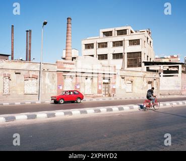 Une scène de rue au canal Mahmoudiya à Alexandrie, Egypte en 2006. L'image montre un bâtiment industriel, une voiture rouge et une personne sur un vélo, capturant la vie quotidienne et l'architecture. Banque D'Images