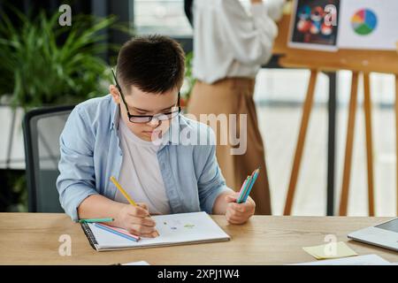 Un garçon atteint du syndrome de Down dessine à un bureau pendant que sa mère travaille en arrière-plan. Banque D'Images