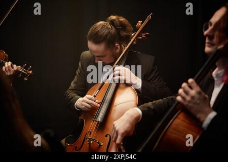 Plan moyen d'un musicien masculin regardant vers le bas, tout en strumming violoncelle jouant de la musique classique avec dévouement dans un ensemble à cordes en studio avec lumière supérieure contrastée sur fond noir Banque D'Images