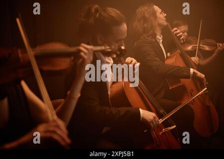 Quatuor à cordes jouant passionnément des violoncelles et des violons tout en étant assis en rangée sur scène dans la lumière orange brumeuse, espace de copie Banque D'Images