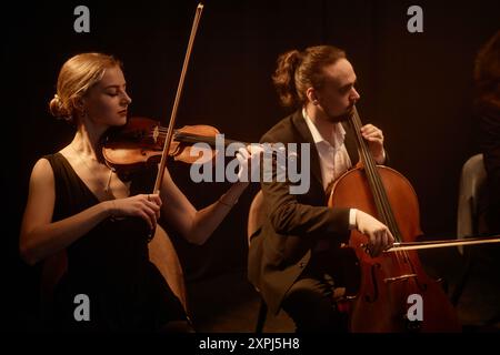 Vue latérale d'artistes de musique classique féminins et masculins jouant du violon et du violoncelle dans un quatuor à cordes, sur fond noir dans un studio d'enregistrement avec une lumière chaude faible Banque D'Images