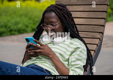Femme noire avec des écouteurs se détend sur un banc en bois à l'extérieur, profitant de son smartphone par une journée ensoleillée dans un parc. Banque D'Images