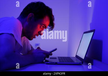 Un homme est concentré sur son smartphone tout en se penchant sur un ordinateur portable dans une pièce éclairée. Les teintes bleues et violettes de l'éclairage créent une atmosphère tamisée Banque D'Images