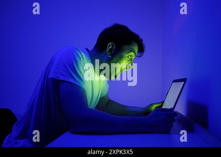 Un homme avec un regard de choc et de surprise est assis dans une pièce faiblement éclairée par la lumière bleue, regardant attentivement un écran. Son expression faciale révèle Banque D'Images