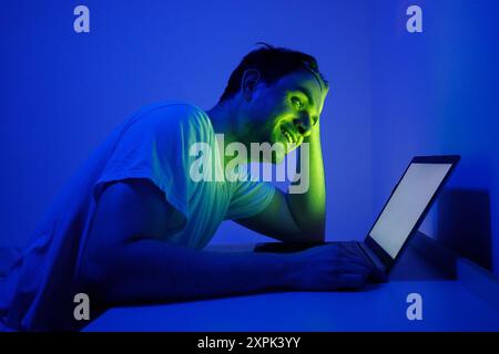 Un jeune homme est assis à un bureau, travaillant sur un ordinateur portable tard dans la nuit. La salle est éclairée par un éclairage bleu et vert, créant une atmosphère vibrante. Lui Banque D'Images