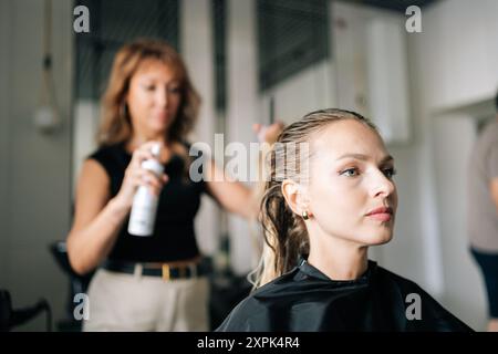 Coiffeur féminin pulvérisant le liquide de mise en place de cheveux sur les cheveux blonds de client de femme. Dame cliente avec de longs cheveux blonds obtenant coiffure élégante fait au salon. Banque D'Images