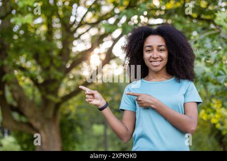 Femme afro-américaine souriante en chemise bleue pointant latéralement dans le cadre du parc avec des arbres en arrière-plan. Concept de bonheur, plein air et vibrations positives. Banque D'Images