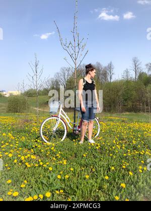 Une fille se tient à côté d'un vélo dans le pré de printemps. Derrière son sac à dos. Il y a beaucoup de pissenlits en fleurs dans le pré. Banque D'Images