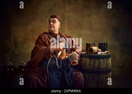 Moine médiéval en vêtements d'époque versant de la bière dans la tasse de la bouilloire, assis dans la cave faiblement éclairée avec tonneau en bois et tasse de bière sur le dessus. Oktoberfest Banque D'Images