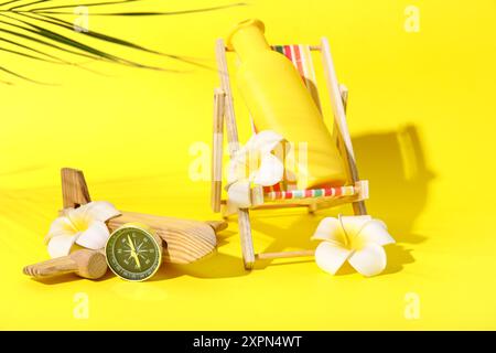 Chaise-terrasse miniature avec bouteille de crème solaire, boussole, avion en bois et fleurs de plumeria sur fond jaune Banque D'Images