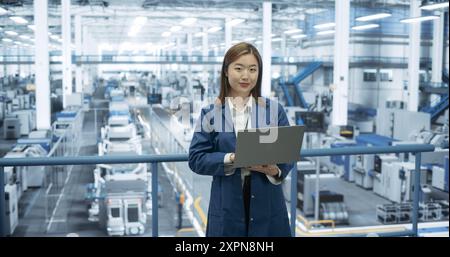 Portrait d'un jeune technicien asiatique utilisant un ordinateur portable et regardant un appareil photo. Spécialiste féminine industrielle portant un manteau bleu, travaillant dans une usine d'électronique moderne Banque D'Images