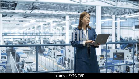Portrait d'un bel ingénieur japonais utilisant un ordinateur portable et regardant autour d'une usine avec un équipement produisant des composants électroniques modernes pour différentes industries Banque D'Images