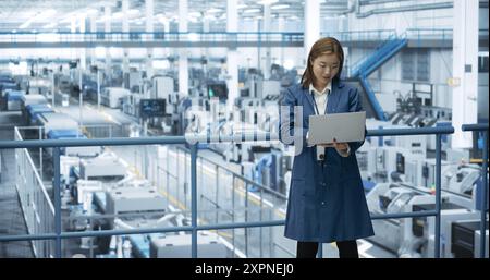 Ingénieur féminin asiatique debout dans une usine, travaillant sur un ordinateur portable, recherchant et développant des technologies pour produire de l'électronique à une fabrication automatisée moderne avec des bras robotiques Banque D'Images