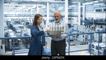 Ingénieur masculin et chef de projet féminin debout sur une plate-forme, parlant, utilisant un ordinateur portable et surveillant la production dans une fabrication moderne d'électronique automatisée avec des robots IA Banque D'Images