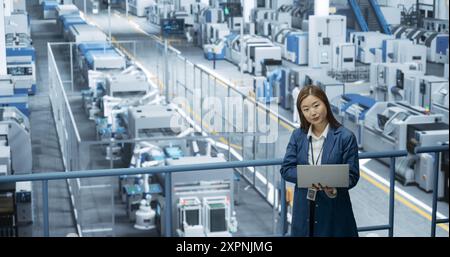 Ingénieur féminin asiatique debout dans une usine, travaillant sur un ordinateur portable, recherchant et développant des technologies pour produire de l'électronique à une fabrication automatisée moderne avec des bras robotiques Banque D'Images