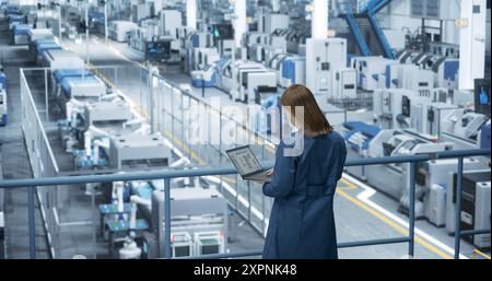 Ingénieur féminin asiatique debout dans une usine, travaillant sur un ordinateur portable, recherchant et développant des technologies pour produire de l'électronique à une fabrication automatisée moderne avec des bras robotiques Banque D'Images