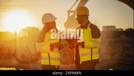 Cliché cinématographique à l'heure d'or du chantier de construction : ingénieur civil masculin caucasien et inspecteur féminin hispanique parlant, à l'aide d'une tablette. Camions, pelles hydrauliques, chargeurs travaillant sur un projet d'aménagement du territoire. Banque D'Images