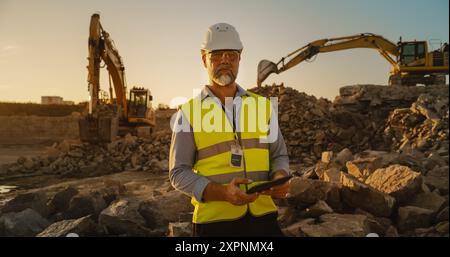 Ingénieur civil barbu à succès portant des lunettes de protection et regardant la caméra sur le chantier de construction un jour ensoleillé alors que des machines lourdes travaillent. Homme tenant la tablette, portant un casque de sécurité, gilet de sécurité. Banque D'Images