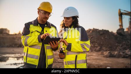 Ingénieur civil masculin caucasien parlant à l'inspecteur féminin hispanique et en utilisant un ordinateur tablette sur le site de construction d'un nouveau bâtiment. Les promoteurs immobiliers discutent d'affaires, les excavatrices travaillent. Banque D'Images