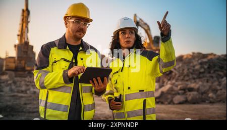 Ingénieur civil masculin caucasien parlant à l'inspecteur féminin hispanique et en utilisant un ordinateur tablette sur le site de construction du nouveau bâtiment. Les promoteurs immobiliers discutent d'affaires, les excavatrices travaillent. Banque D'Images