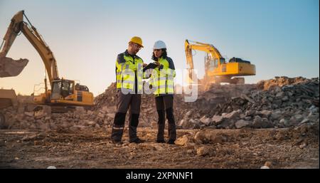 Ingénieur civil masculin caucasien parlant à l'inspecteur féminin hispanique et en utilisant la tablette sur le site de construction du nouveau complexe d'appartements. Les promoteurs immobiliers discutent d'affaires, les excavatrices travaillent. Banque D'Images