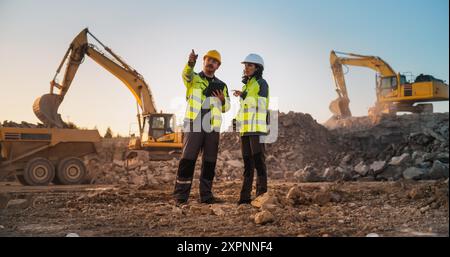 Ingénieur civil masculin caucasien parlant à l'inspecteur féminin hispanique et en utilisant la tablette sur le site de construction du complexe d'appartements. Les promoteurs immobiliers discutent d'affaires, les excavatrices travaillent. Banque D'Images