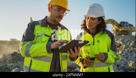 Ingénieur civil masculin caucasien parlant à l'inspecteur féminin hispanique et en utilisant la tablette sur le site de construction du nouveau bâtiment résidentiel. Les promoteurs immobiliers discutent d'affaires, les excavatrices travaillent. Banque D'Images
