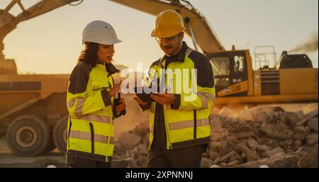 Cliché cinématographique à l'heure d'or du chantier de construction : ingénieur civil masculin caucasien et inspecteur féminin hispanique parlant, à l'aide d'une tablette. Camions et pelles hydrauliques travaillant sur le projet d'aménagement du territoire. Banque D'Images