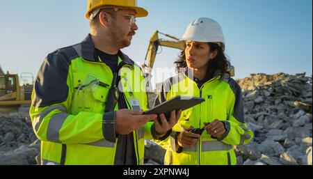 Ingénieur civil masculin caucasien parlant à l'inspecteur féminin hispanique et en utilisant la tablette sur le site de construction de l'immeuble d'appartements. Les promoteurs immobiliers discutent d'affaires, les excavatrices travaillent. Banque D'Images