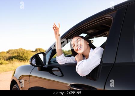 Jeune femme souriante penchée par la fenêtre de la voiture, vue de l'extérieur. J'apprécie le voyage Banque D'Images