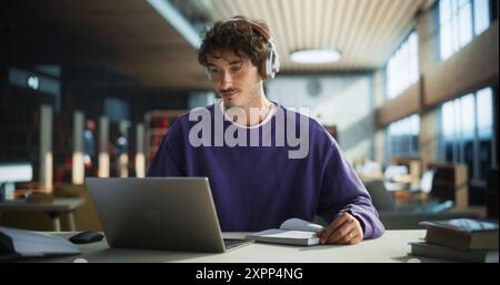 Beau étudiant écrivant un essai sur ordinateur portable, réfléchissant au sujet, brainstorming, trouver des solutions. Jeune homme élégant étudiant dans une bibliothèque publique, écoutant lecture dans des écouteurs Banque D'Images