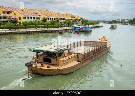 La barge se déplace sur la rivière. Une barge flottante sur une rivière Thu bon à Hoi an Vietnam. Ancienne ville, maisons colorées, conteneurs d'expédition sur barge fluviale. Banque D'Images