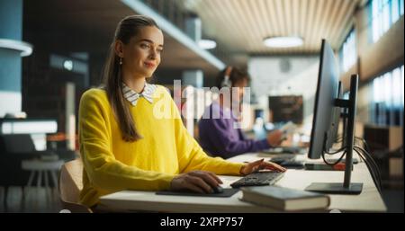 Belle étudiante féminine caucasienne en jaune Jumper travaillant sur sa thèse d'école sur un ordinateur de bureau. Jeune femme intelligente étudiant en ligne dans une bibliothèque publique tranquille avec des étagères de livres Banque D'Images