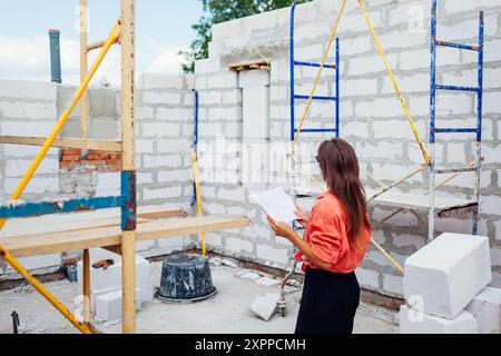 Vue arrière de la jeune femme vérifiant sa maison en construction debout à l'intérieur du bâtiment avec plan de plan. Le futur propriétaire imagine une maison confortable Banque D'Images