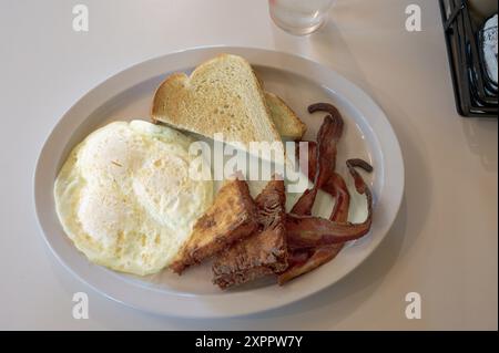 Petit déjeuner traditionnel composé d'œufs, bacon, pain grillé et gâteaux de pommes de terre frits sur une assiette de couleur blanche Banque D'Images