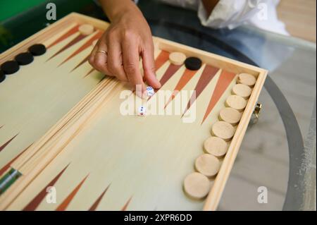 Gros plan d'une main qui roule des dés sur un plateau de backgammon en bois, présentant la stratégie et le divertissement dans une configuration de jeu de table classique. Banque D'Images