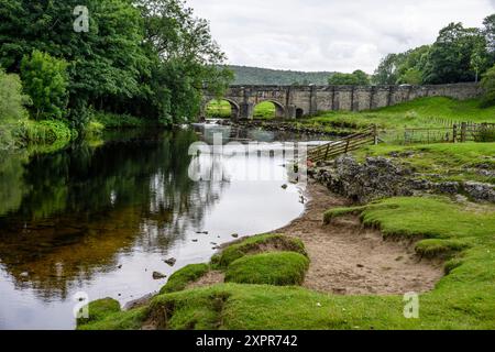 Un pont de campagne serein enjambant une rivière calme entouré de verdure luxuriante et de rives rocheuses. Banque D'Images