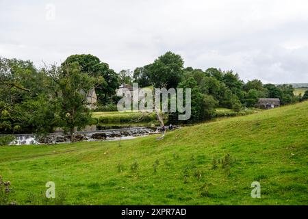 Grassington Yorkshire UK-27 juillet 2024. Scène de campagne tranquille avec une maison en pierre historique au bord d'une rivière entourée d'une végétation luxuriante. Banque D'Images