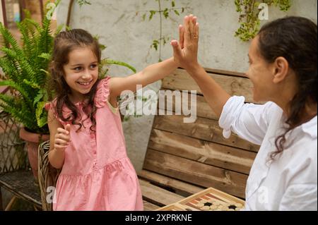 Moment joyeux entre une jeune fille et sa mère alors qu'ils High-Five lors d'un jeu de société dans un jardin extérieur. Liens familiaux et amusement en plein air. Banque D'Images