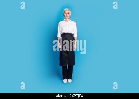 Photo du corps entier de la femme senior attrayante propriétaire de petit café d'entreprise debout habillé uniforme de serveuse isolé sur fond de couleur bleue Banque D'Images